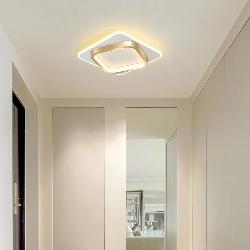 Gold-accented square LED ceiling light fixture in a modern, minimalist hallway with soft white walls and a full-length mirror.